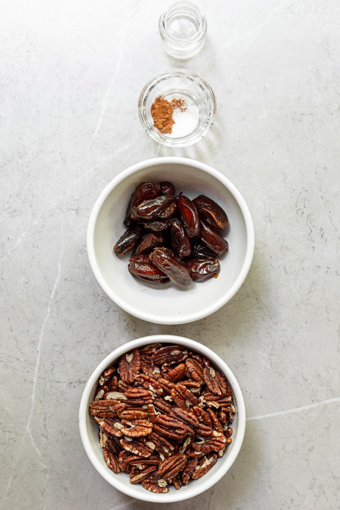 4 bowls on a marble counter with the ingredients for the no-bake crust - pecans, dates, cinnamon/salt, and vanilla extract