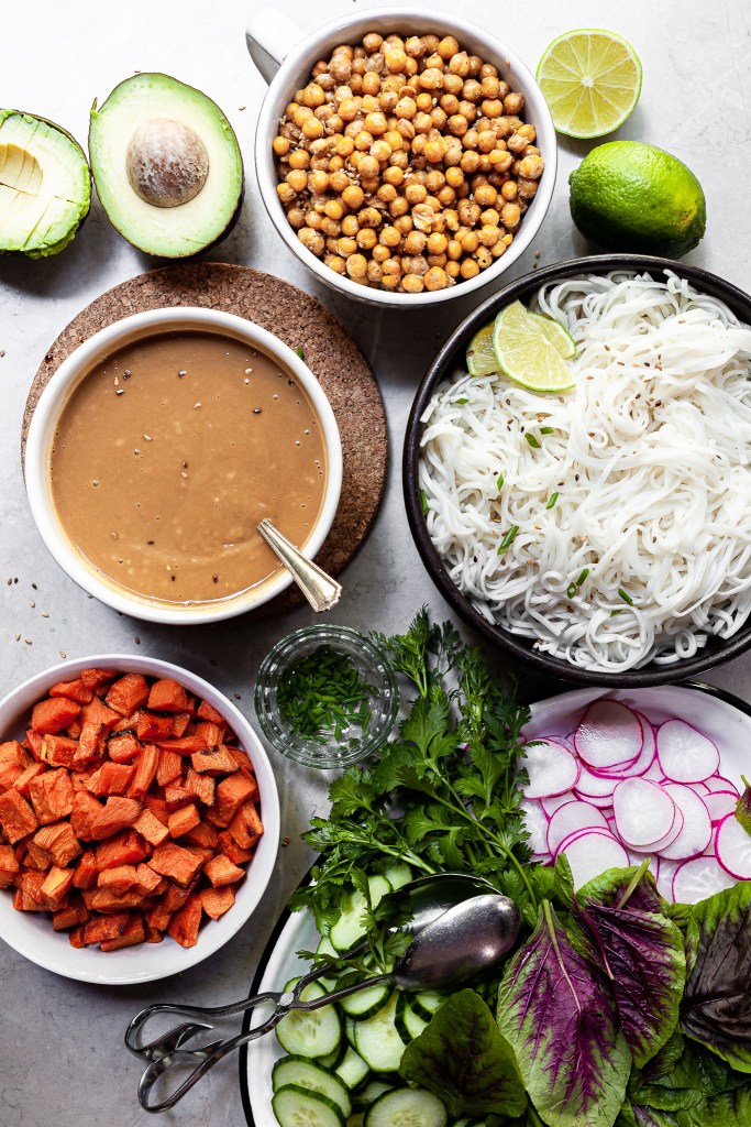 All of the components for this recipe laid out in bowls: roasted sweet potatoes, roasted chickpeas, rice noodles, fresh vegetables and sesame ginger sauce