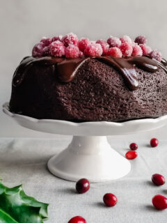Vegan Cranberry Chocolate Fudge Cake with Ganache and Sugared Cranberries served on a white platter with holly and cranberries on the table