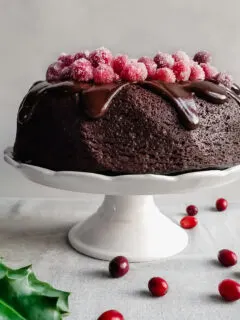Vegan Cranberry Chocolate Fudge Cake with Ganache and Sugared Cranberries served on a white platter with holly and cranberries on the table