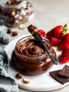 Jar filled with hazelnut chocolate spread beside fresh berries and a jar of whole hazelnuts in the background
