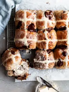 Flatlay of hot cross buns on a cooling rack, with one having been cut in half and smeared with vegan butter