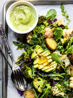 A serving of kale and roasted vegetable salad on an aluminium tray served with a small white bowl filled with creamy light green avocado Caesar dressing