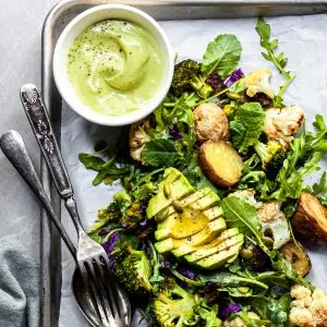A serving of kale and roasted vegetable salad on an aluminium tray served with a small white bowl filled with creamy light green avocado Caesar dressing