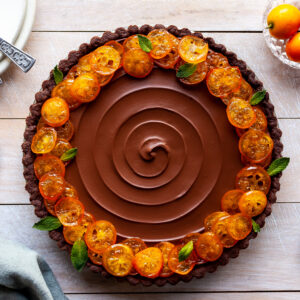 Vegan Chocolate Orange Tart with Candied Kumquats sits beside serving plates and fresh kumquats