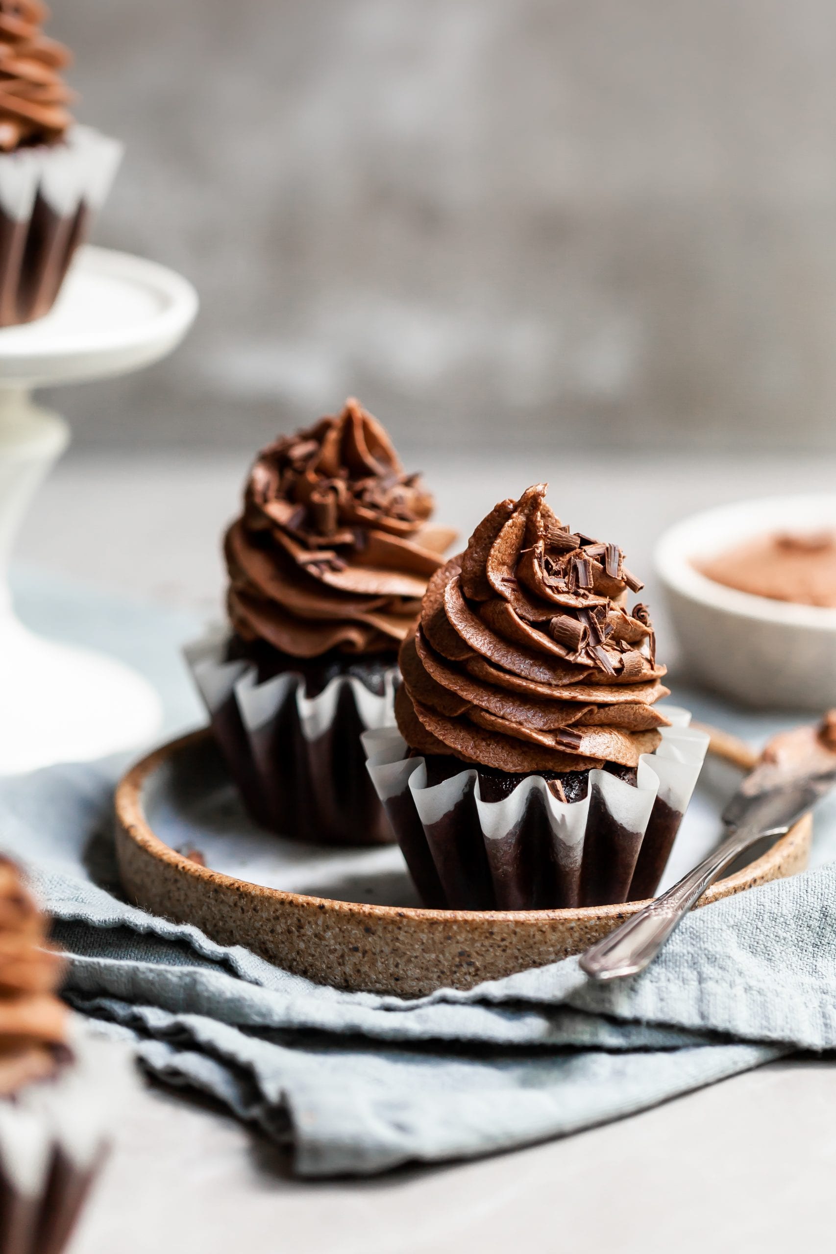 Two chocolate cupcakes on a plate with a bowl of fluffy chocolate ganache frosting on the side