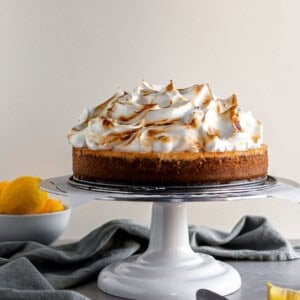Lemon Meringue Cheesecake sitting on a white cake stand beside a bowl of lemons