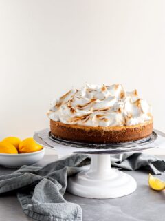 Vegan Lemon Meringue Cheesecake on a white cake stand beside a bowl of lemons