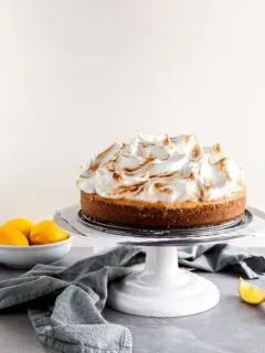 Vegan Lemon Meringue Cheesecake on a white cake stand beside a bowl of lemons