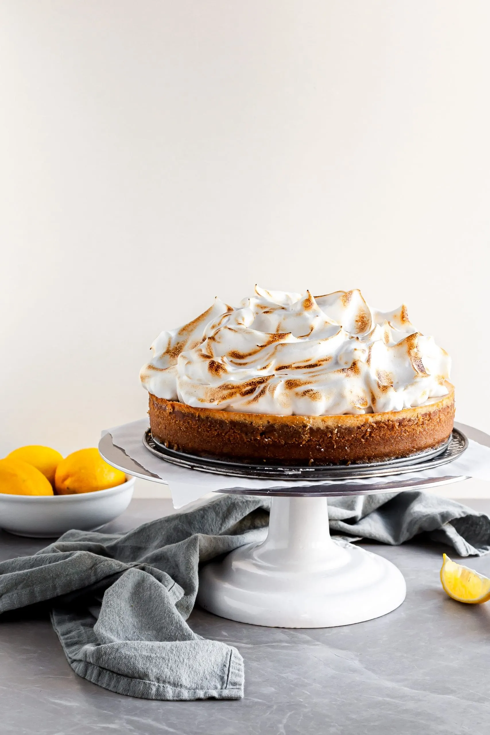 Vegan Lemon Meringue Cheesecake on a white cake stand beside a bowl of lemons