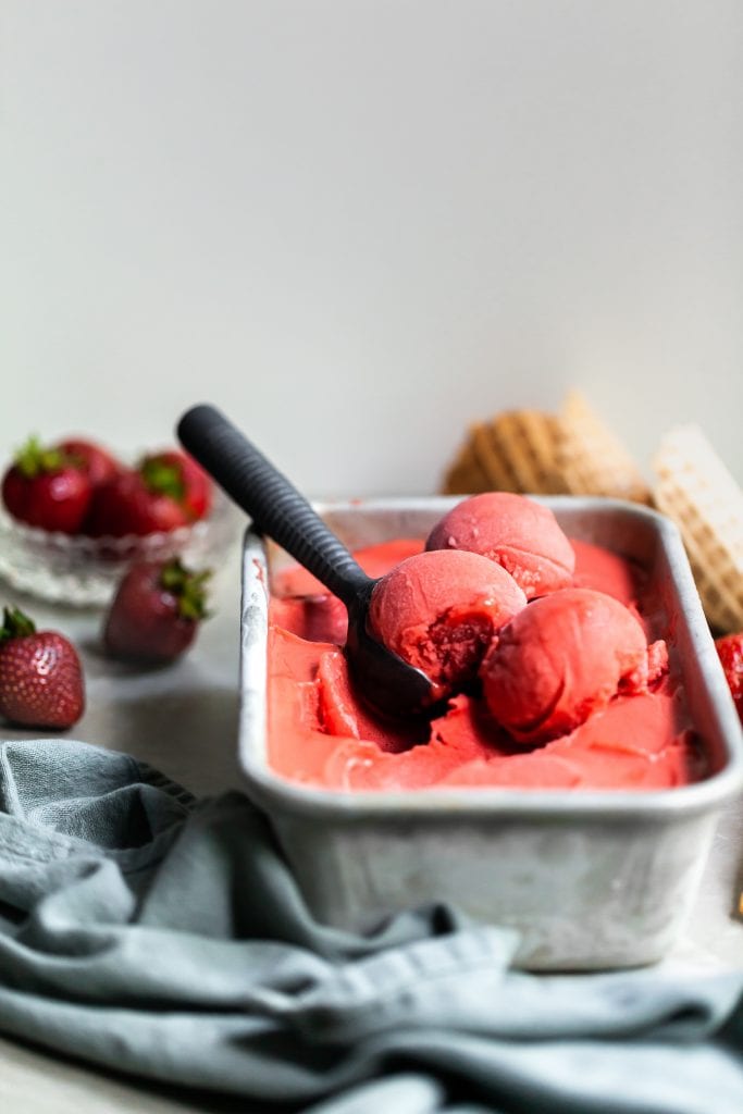 Side view of a loaf pan of strawberry rhubarb sorbet with 3 scoops ready to be served, beside some cones and fresh strawberries