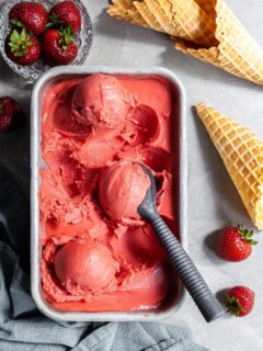 Flatlay of strawberry rhubarb sorbet scoops in a rectangular pan beside a dish of fresh strabwerries and some ice cream cones