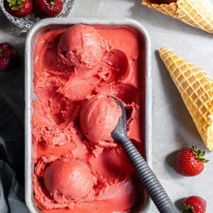 Flatlay of strawberry rhubarb sorbet scoops in a rectangular pan beside a dish of fresh strabwerries and some ice cream cones