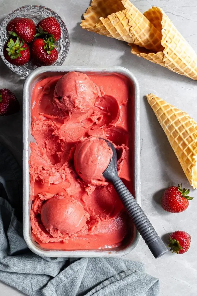 Flatlay of strawberry rhubarb sorbet scoops in a rectangular pan beside a dish of fresh strabwerries and some ice cream cones