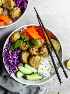 White bowl and chopsticks with rice noodles, chcikpea balls covered in yellow curry sauce, and raw veggies