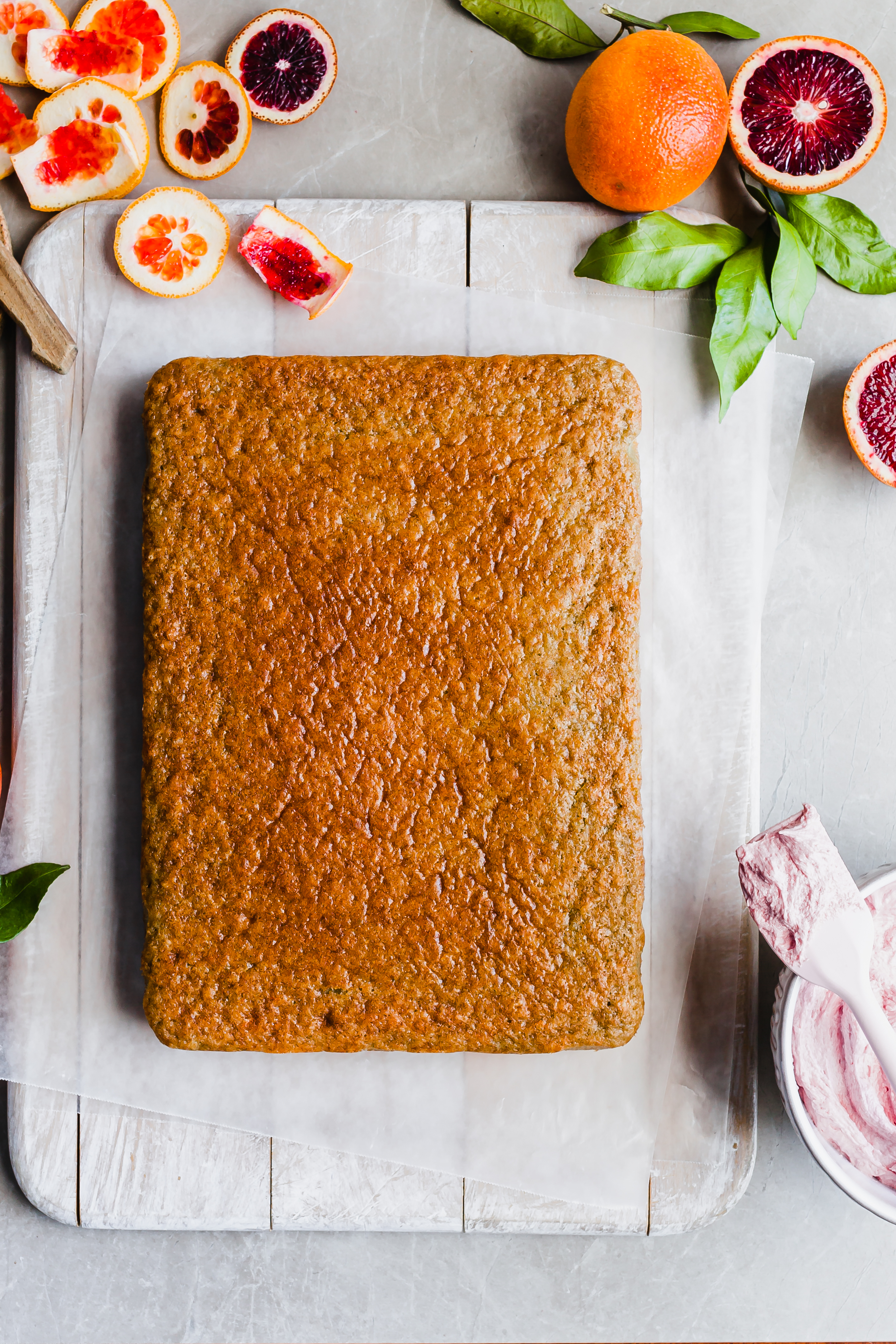 A sheet cake lies on a white cutting board beside a bowl of pink frosting, surrounded by cut blood oranges and rind