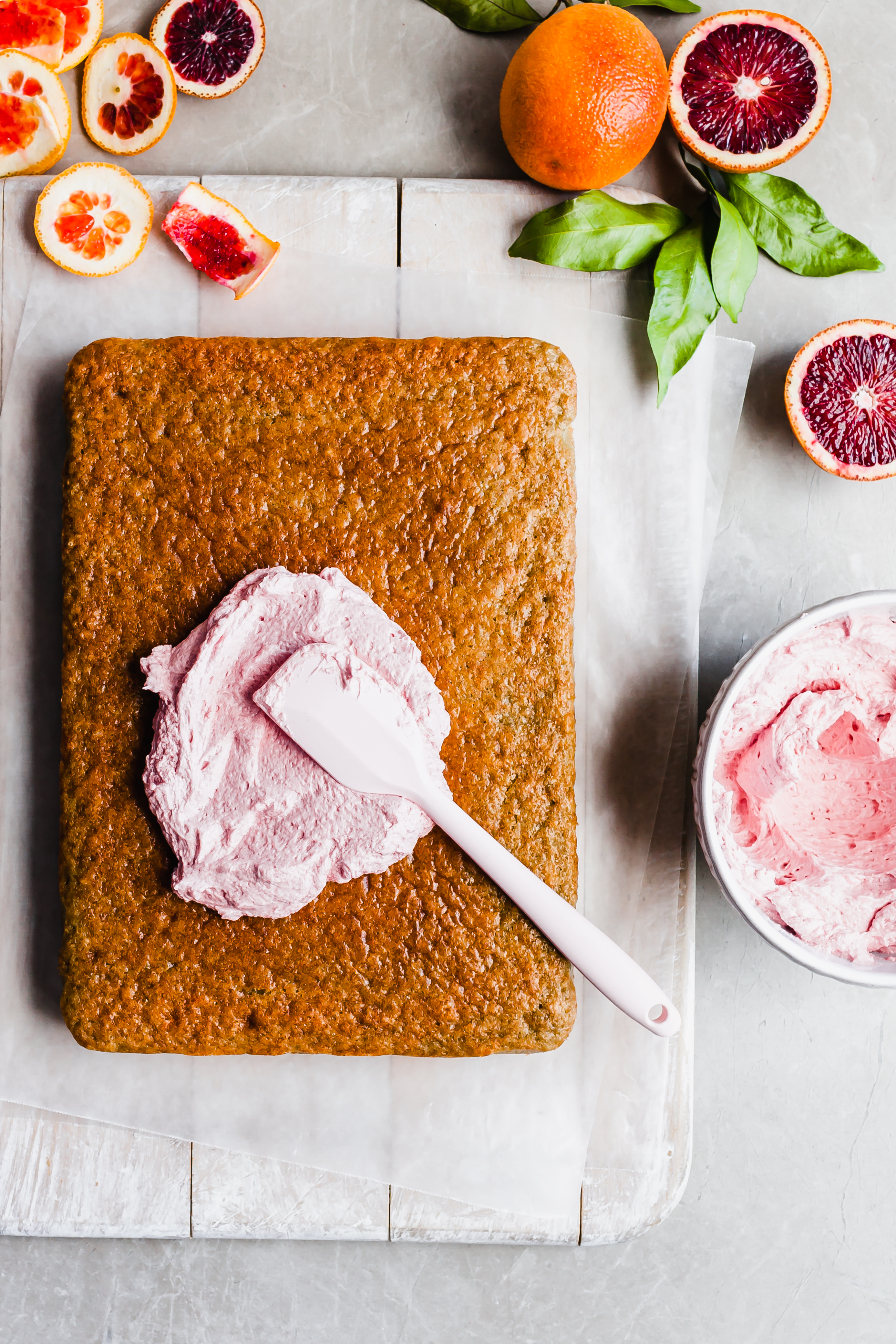 A sheet cake lies on a white cutting board with the first dollop of pink frosting applied and the spatula left on top. Beside it is a bowl of pink frosting and sliced blood oranges