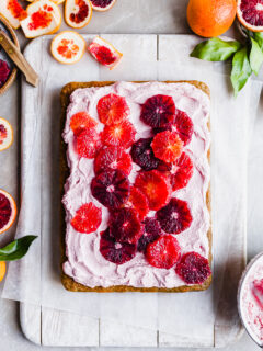Flatlay of a sheet cake covered in pink frosting and slices of blood oranges, the background littered with orange rinds
