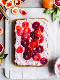 Flatlay of a sheet cake covered in pink frosting and slices of blood oranges, the background littered with orange rinds