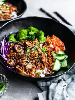 Angled view of a black bowl filled with rice topped with lentils, shiitakes, broccoli and kimchi
