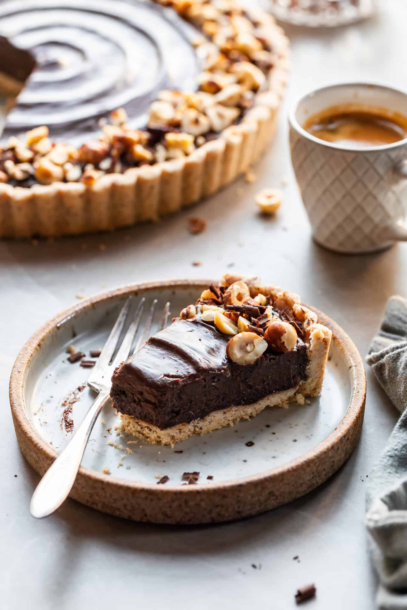 Close up of a slice of vegan tart with a bite taken, and a small espresso in the background beside the chocolate hazelnut tart