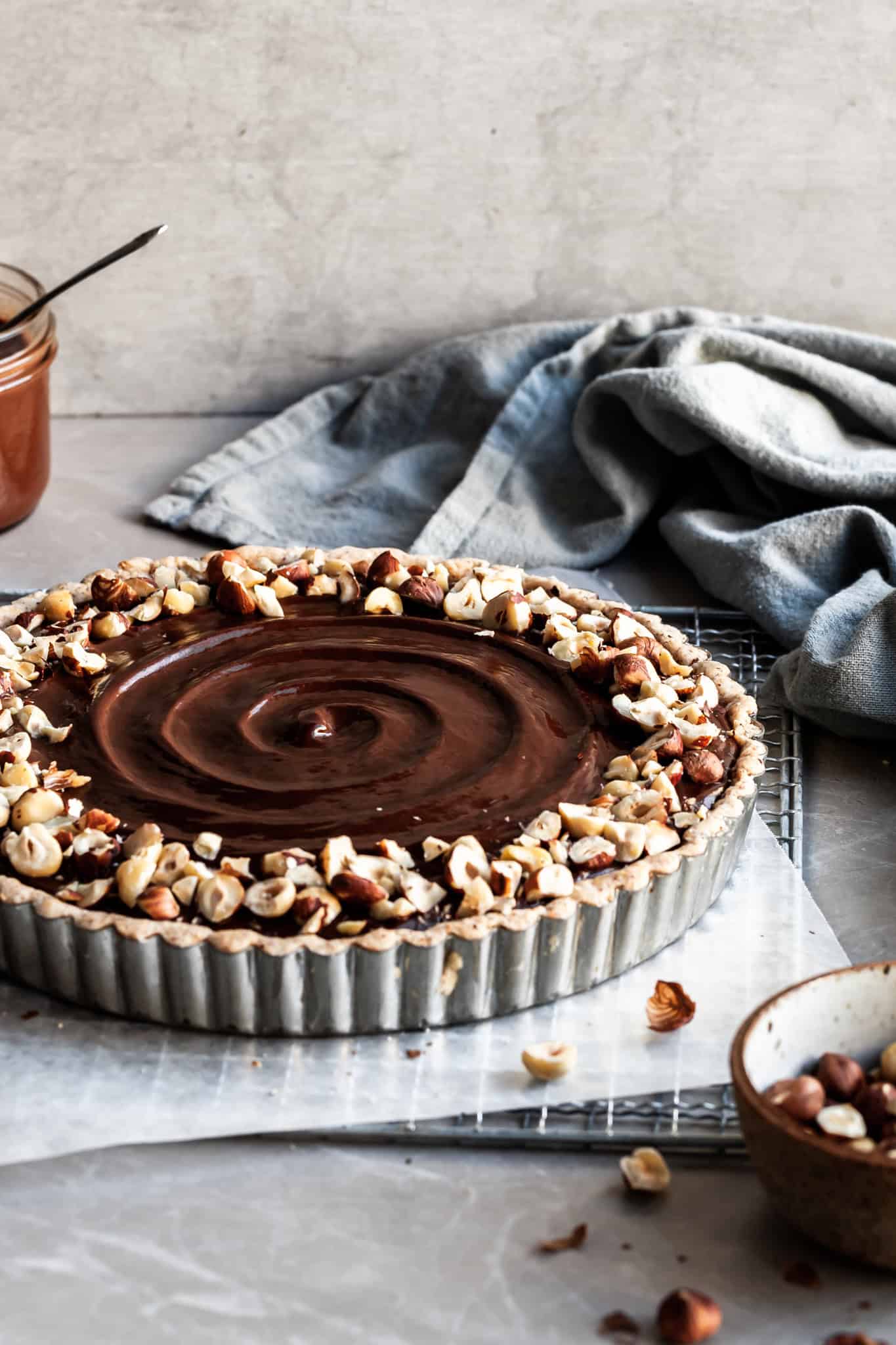 side view chocolate hazelnut tart with a small bowl of hazelnuts in the foreground and a jar of chocolate hazelnut spread in the background