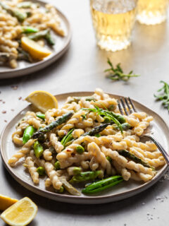 Side view of a plate of gemelli pasta in a light cream sauce with asparagus and spring peas with glasses of wine in the background