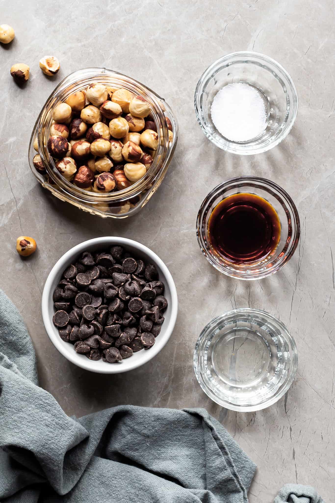 Ingredients measured out for the homemade nutella in small glass bowls: chocolate chips, hazelnuts, vanilla, salt and coconut oil