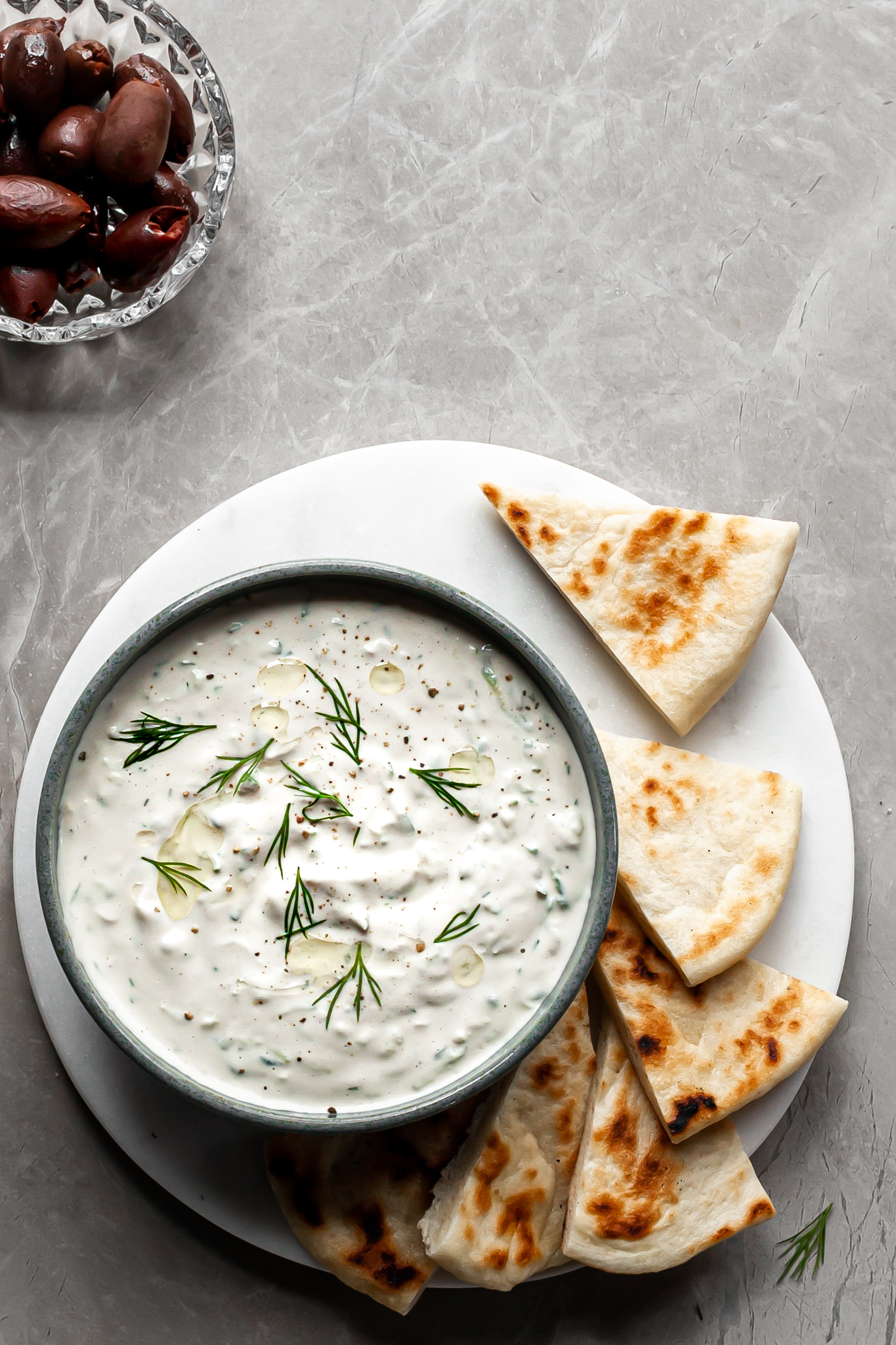 View looking down on a bowl of vegan tzatziki served with flatbread, cucumber slices and black olives