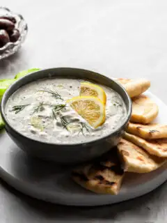 Grey bowl of tzatziki sauce garnished with lemon wedges, served with greek flatbread and slices of cucumber