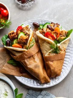 2 cauliflower gyro wraps wrapped in brown paper on a plate beside a bowl of tzatziki
