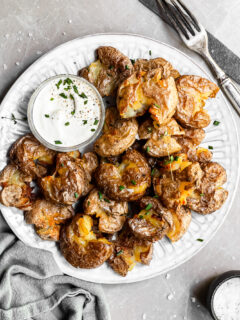 White plate filled with crispy smashed potatoes and a small glass bowl of vegan sour cream
