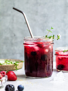 Jar filled with vibrant purplish reddish fizzy drink, garnished with thyme sprigs and served with a straw.