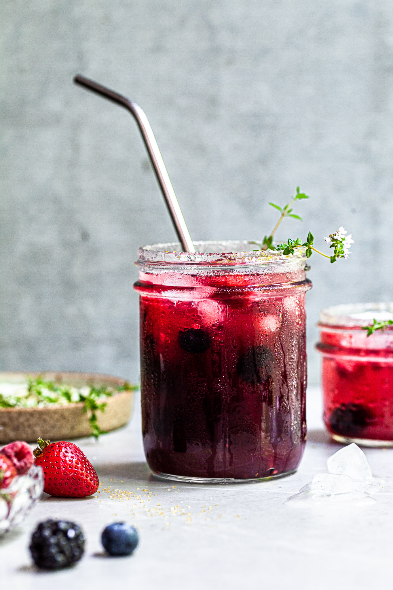 Jar filled with vibrant purplish reddish fizzy drink, garnished with thyme sprigs and served with a straw.