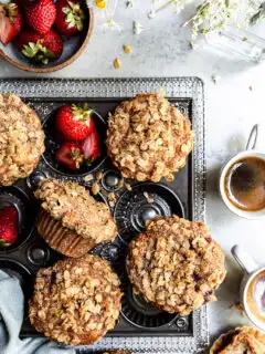 View looking down on a baking tray of muffins beside a bowl of strawberries, a vase of wildflowers and 2 some cups of espresso