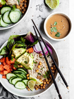 white bowl filled with rice noodles, cubes of sweet potatoes, chickpeas and cucumbers, with a small bowl of tahini dressing on the side