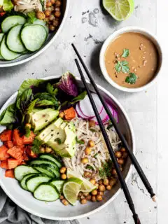 white bowl filled with rice noodles, cubes of sweet potatoes, chickpeas and cucumbers, with a small bowl of tahini dressing on the side