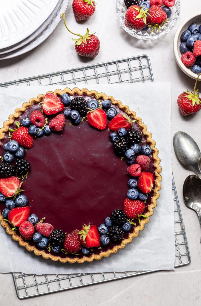 flatlay of a purple berry tart, garnished with summer berries, and several small bowls with berries beside it