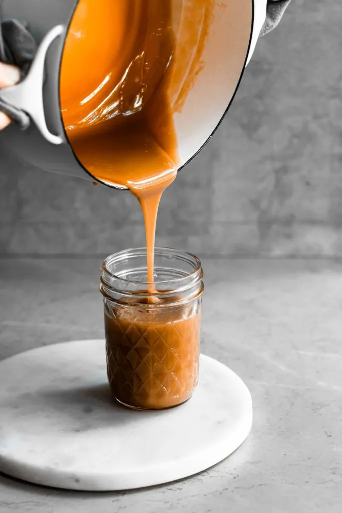 caramel being poured from a white pot into a clear glass jar