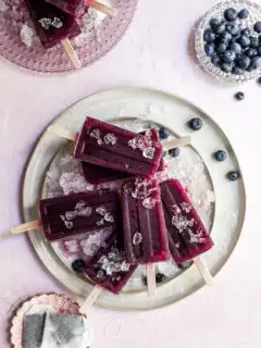 a cream colored plate covered in crushed ice with purple popsicles and blueberries resting on top