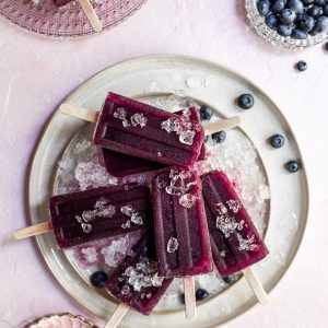 a cream colored plate covered in crushed ice with purple popsicles and blueberries resting on top