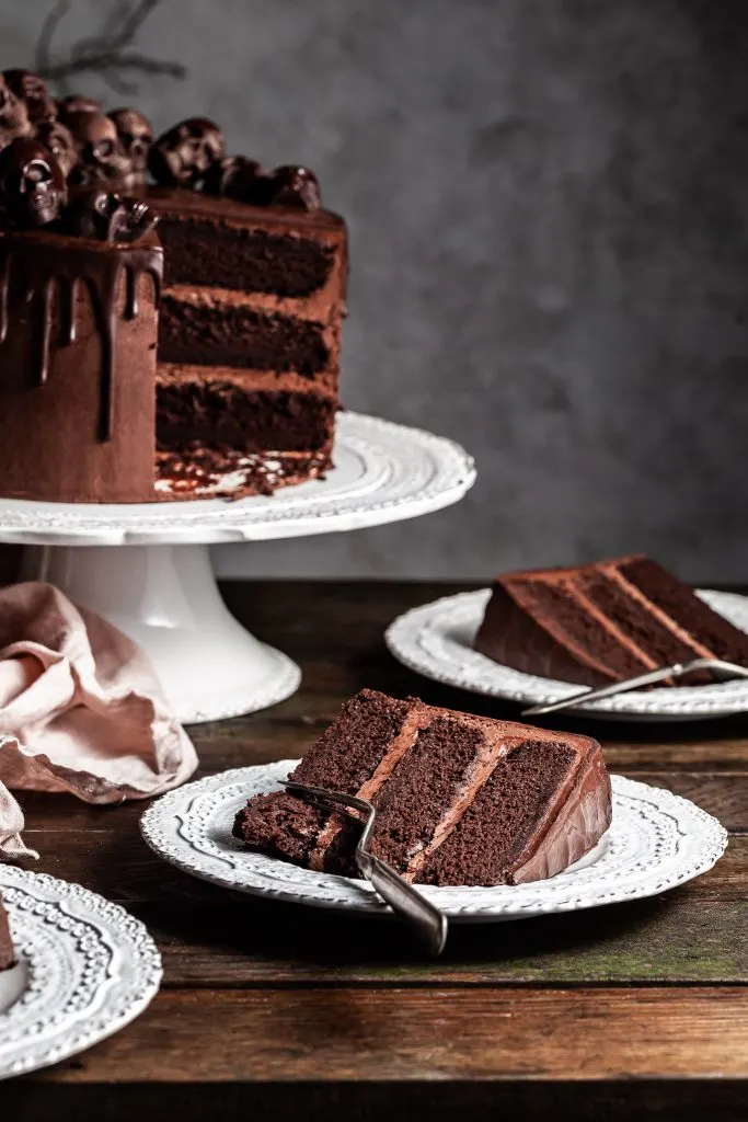 A slice of chocolate cake on a white plate, with another slice lies on a plate in the background beside a white pedestal cake stand holding the cut triple layer chocolate Halloween themed cake