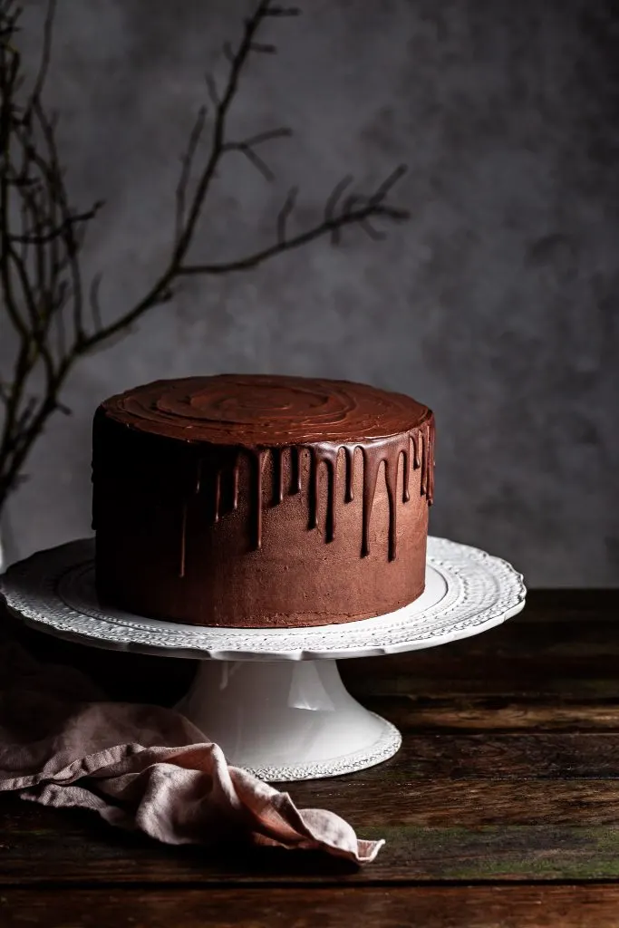 Vegan chocolate layer drip cake on a white cake stand with a branch in the dark background