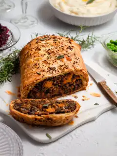 A vegan Wellington on a white marble platter sitting on a tabelscape with a bowl of whipped potatoes, green peas, cranberry sauce and wine glasses.