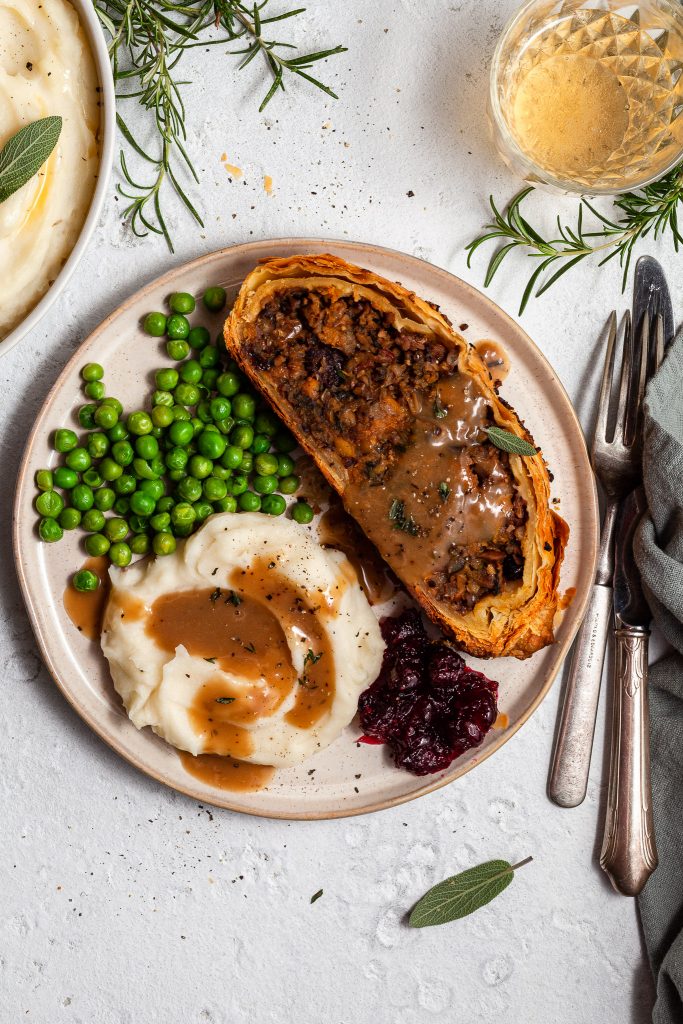 A slice of vegan Wellington on a plate with mashed potatoes, green peas, cranberry sauce and gravy.