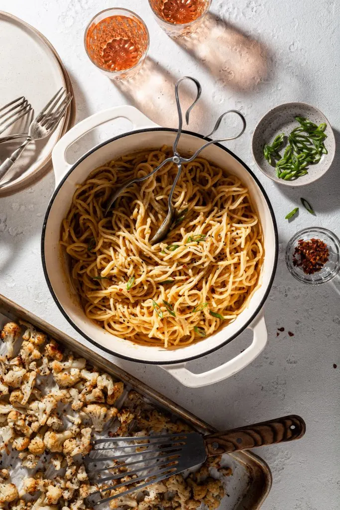 View of a white enamel Dutch oven pot filled with vegan garlic miso spaghetti noodles garnished with sesame seeds and slices of green onions. A baking sheet rests beside the Dutch oven, filled with roasted cauliflower. 