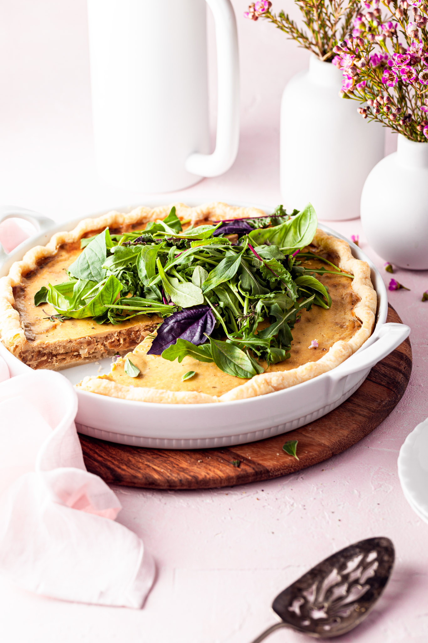 A quiche in a white tart dish with a slice served. White vases are in the background on this brunch table.