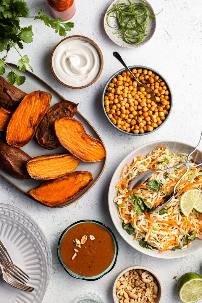 View looking down on the elements of this loaded sweet potato recipe before the sweet potatoes are plated - a platter of baked sweet potatoes, a bowl of coleslaw, a bowl of peanut sauce, a bowl of crispy chickpeas and various garnishes. 