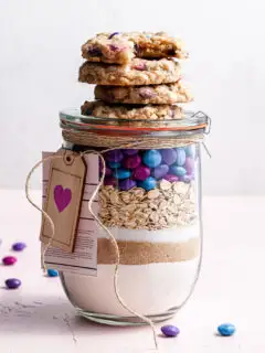 A jar filled with cookie mix, topped with baked vegan m&m cookies.