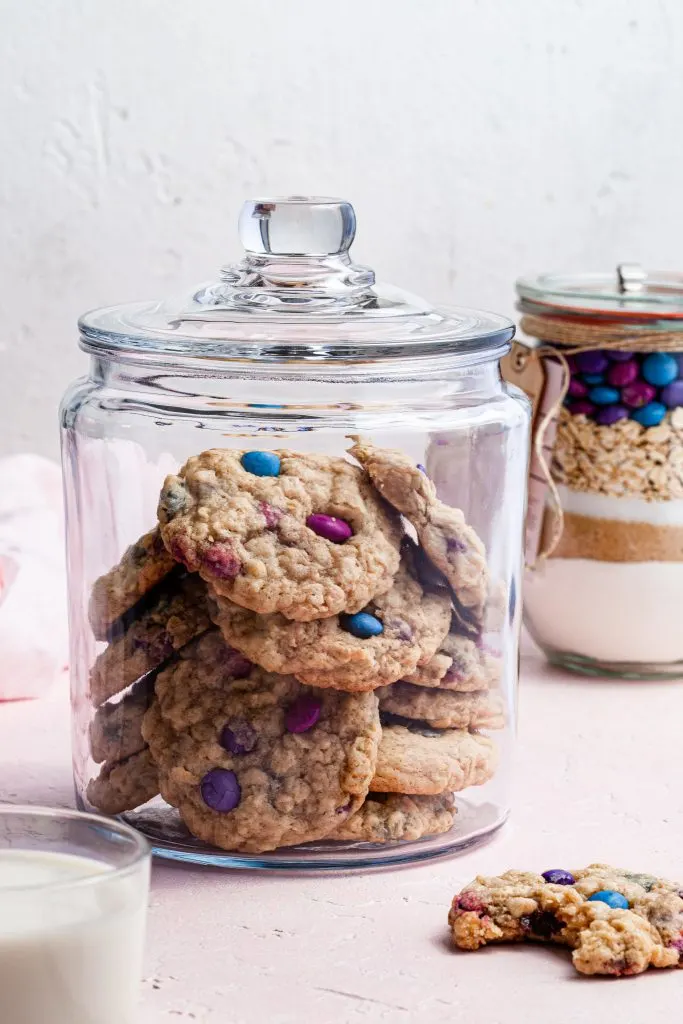 A glass cookie jar filled with cookies, one cookie sits on the counter with a bite missing and a cookie kit jar in the background. 